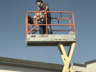 Father and daughter standing on a raised scissor lift platform, preparing balloons together under a clear blue sky.